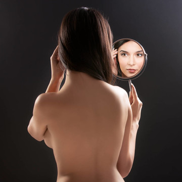 一個(gè)美麗的工作室的照片，一個(gè)年輕女子在鏡子里，而她的背轉(zhuǎn)向相機(jī)。鏡子放在她的左肩上，折射出她笑容。