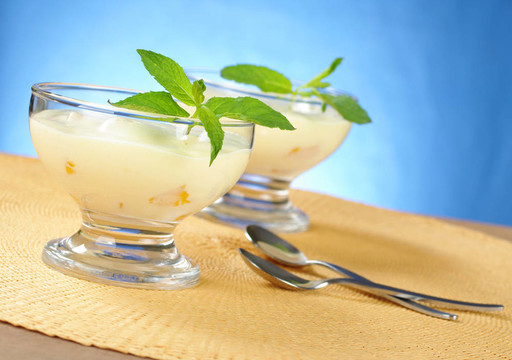芒果奶油奶酪甜點(diǎn)在玻璃碗與甜點(diǎn)匙在黃色表墊藍(lán)色背景（選擇性的重點(diǎn)，重點(diǎn)放在前面的玻璃和葉子）