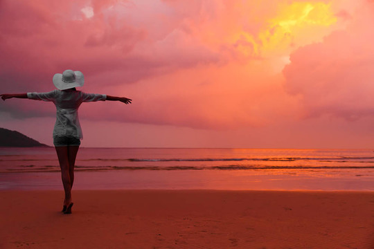 年輕女子戴著帽子站在潮濕的沙灘上望著夕陽紅色的天空