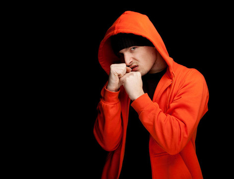 憤怒的年輕人站在橙色運(yùn)動(dòng)衫罩和黑帽；抬起手臂；黑色背景