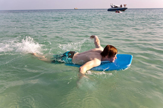 男孩冲浪在美丽的海边的一个小的冲浪板与清澈的水和蓝色的天空