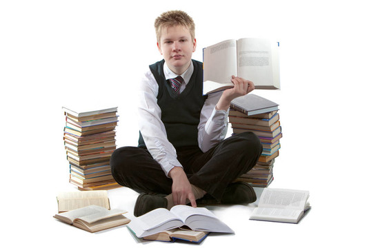 穿著校服的男生坐在地板上；靠近一包書；手里拿著打開的書。