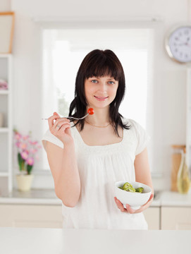 可愛(ài)的黑發(fā)女性吃櫻桃番茄在廚房里端著一碗蔬菜