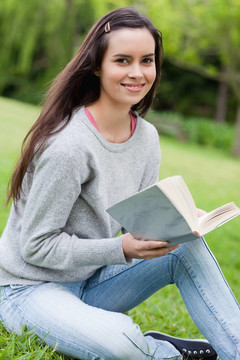 年輕的微笑女孩抱著一本書，坐在草地上，看著相機(jī)