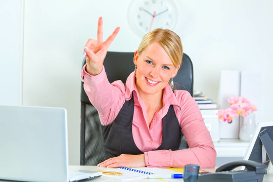 微笑的现代商务女性坐在办公桌前展示胜利姿态