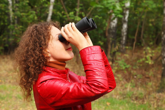 漂亮的年轻女子穿着红色皮夹克；用双筒望远镜向外看