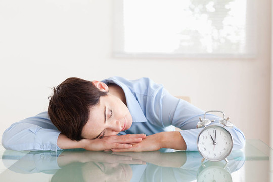 睡眠短頭發(fā)的女人，她的頭靠在桌子旁邊的辦公室里的鬧鐘