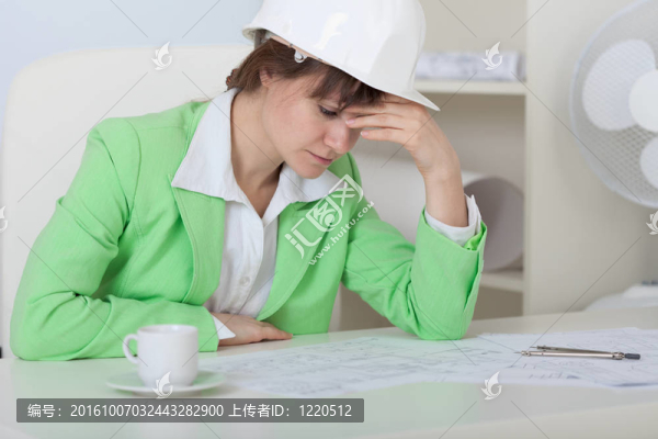 疲倦的女人--戴頭盔的工程師坐在辦公室的桌子上