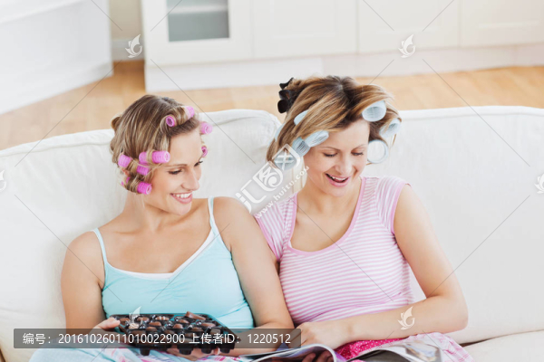 關閉女性朋友與頭發(fā)輥吃巧克力閱讀雜志在家里的沙發(fā)上