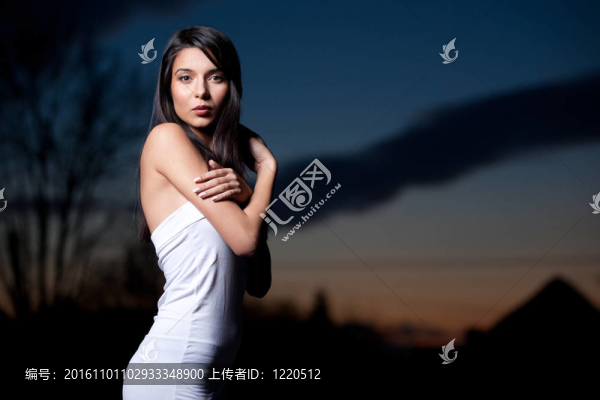 一幅年輕的肖像；美麗的女人；在外面拍攝；在日落的背景。她戴著一頂白色的上衣；她用雙手握住她的肩膀；而