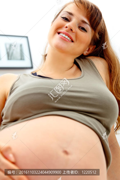 微笑美麗懷孕的女性坐在沙發(fā)上，在家里抱著她的肚子。特寫(xiě)。