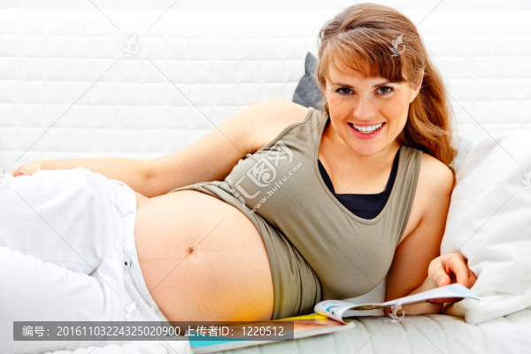微笑美女孕婦在沙發(fā)上與雜志在家放松。