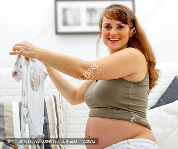 微笑美女懷孕女性坐在沙發(fā)上帶著嬰兒衣服在家。