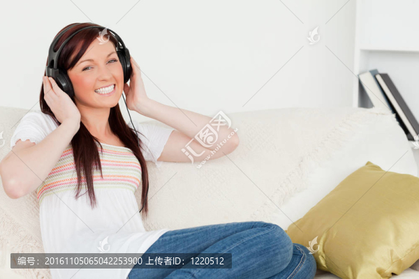 迷人的紅發(fā)女人一邊坐在客廳的沙發(fā)上一邊聽(tīng)音樂(lè)一邊戴著耳機(jī)