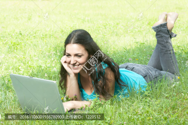 一個可愛的年輕女性躺在公園的草地上用筆記本電腦的肖像