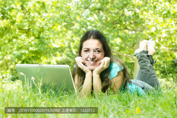 一個可愛的年輕女性躺在公園的草地上用筆記本電腦的肖像