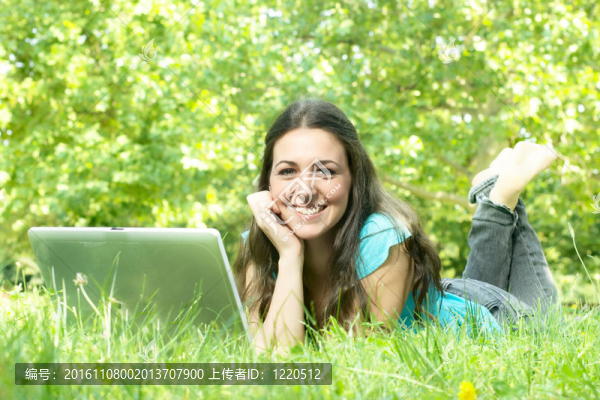 一個可愛的年輕女性躺在公園的草地上用筆記本電腦的肖像