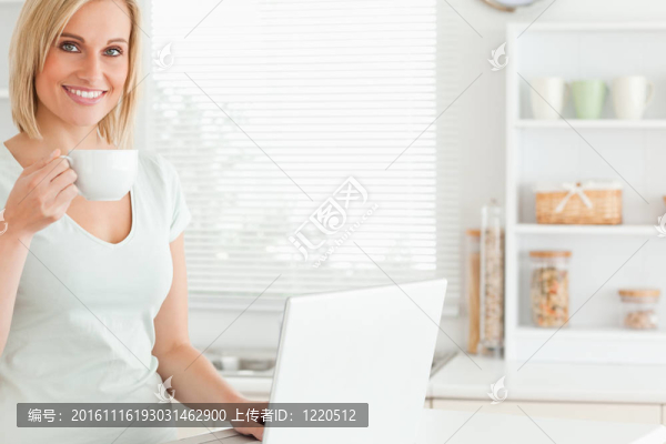 微笑的女人拿著一杯咖啡和一臺(tái)筆記本在廚房里看著相機(jī)