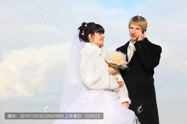 快樂新郎和新娘在冬天擁抱花束在戶外；男人說話電話和女人看男性