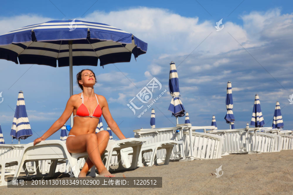 在白色泳衣坐在躺椅里年輕美麗的女子和日光?。话咨梢魏退{(lán)色的雨傘排