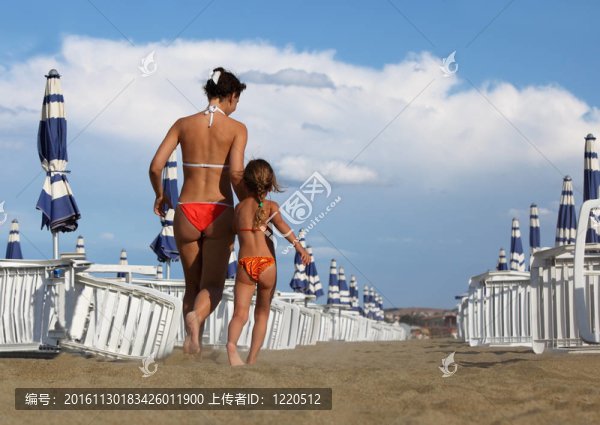 年輕的母親和小女兒穿著泳衣去海灘。母親抱著女兒的手；白色的躺椅和藍(lán)色的雨傘排