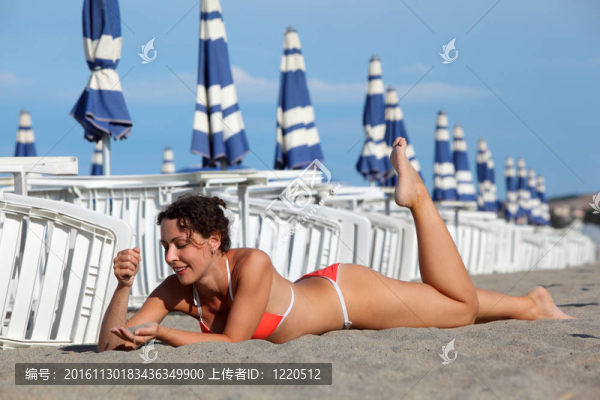 美麗的年輕女子躺在沙灘上曬日光浴海灘。在白色躺椅和藍色的雨傘背景行