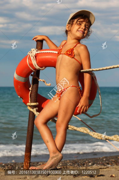 美麗的小女孩穿著泳衣和帽子站在海灘上。她緊貼著金屬桿桿上掛紅色救生圈；