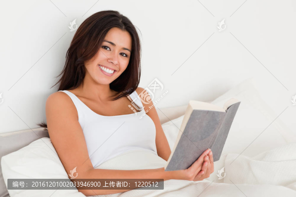 面帶微笑的女人拿著書躺在床上看著相機