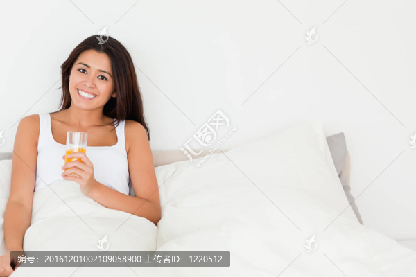 微笑的女人拿著橙汁坐在床上看著臥室里的相機