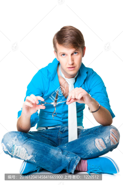 年輕漂亮的男理發(fā)師穿著藍(lán)色襯衫和白色領(lǐng)帶夾剪刀坐在孤立的白色背景