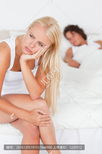 一個(gè)心煩意亂的女人坐在床上的肖像，而她的未婚夫正在睡覺(jué)的相機(jī)專注于她