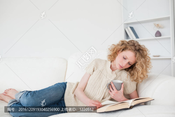 漂亮的金發(fā)女人讀一本書，捧著一杯咖啡，躺在她公寓的沙發(fā)上
