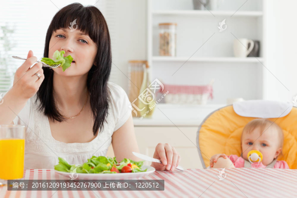 漂亮的黑發(fā)女人吃沙拉在她旁邊的寶寶坐在廚房