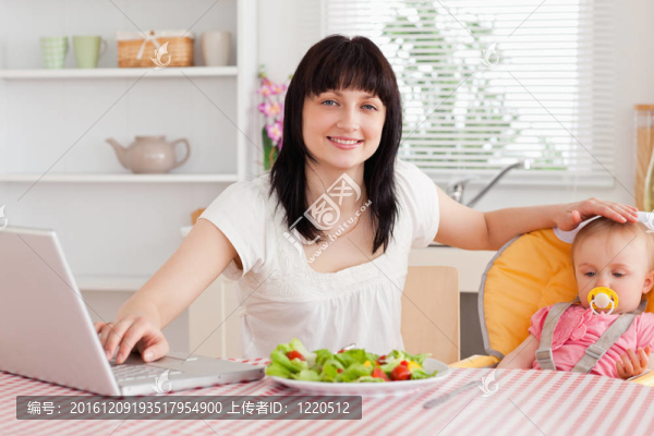 漂亮的黑發(fā)女人吃沙拉在她的嬰兒而輕松的在廚房里用她的筆記本電腦