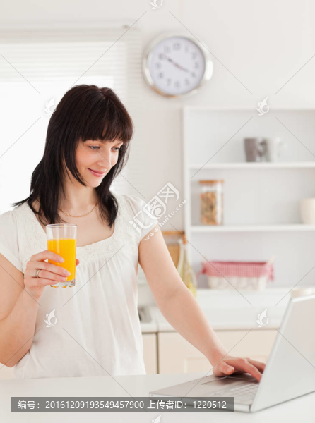 漂亮的黑發(fā)女人拿著一杯橙汁而輕松的在廚房里用她的筆記本電腦