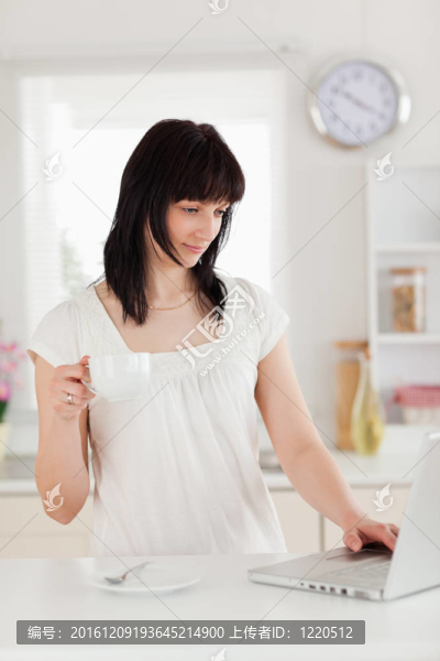 漂亮的黑發(fā)女人端著一杯咖啡而輕松的在廚房里用她的筆記本電腦