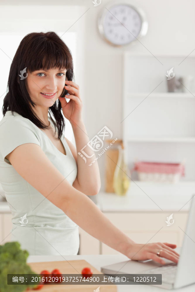 可愛的黑發(fā)女子在電話中放松的同時(shí)在廚房里用她的筆記本電腦