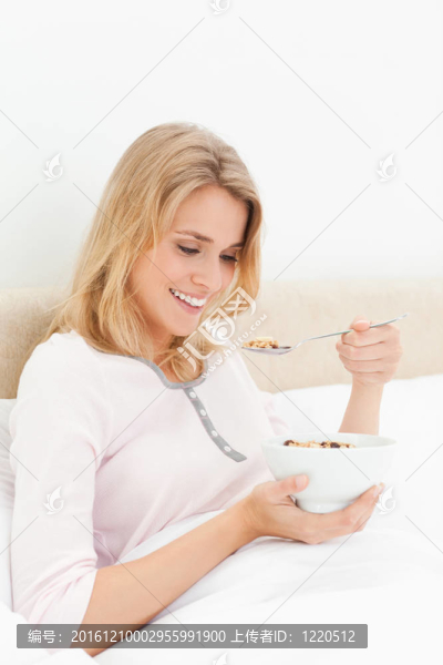 一個(gè)女人坐在床上，手里拿著一碗麥片粥，一邊看著它，一邊微笑著，嘴里放著一勺麥片粥。