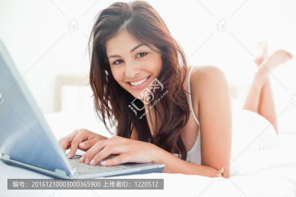 一個面帶微笑的女人躺在床上，手里拿著筆記本電腦；放松著，交叉著她抬起的腿。