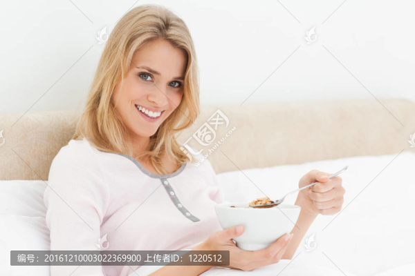一個女人坐在床上；微笑著拿著一個碗和一個稍微舉起的麥片粥。