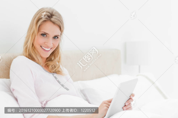 一個(gè)女人坐在床上用她的平板電腦；一邊看著一邊微笑。