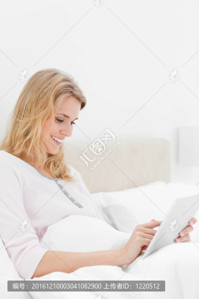 一個女人坐在床上，一邊看著一邊微笑著看的平板電腦。
