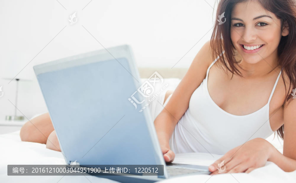 一名女子躺在床上，她與她的筆記本電腦互動，而直向前看和微笑。