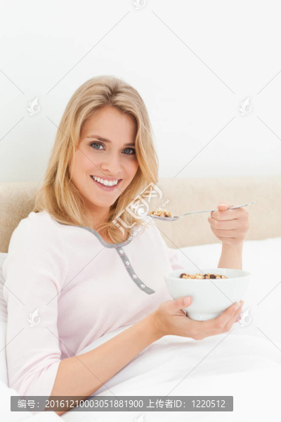 一個(gè)女人坐在床上，手里拿著一碗麥片粥，嘴邊放著一勺麥片粥，一邊向前看，一邊微笑著。