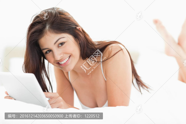 一個女人躺在床上，手里拿著一臺平板電腦，雙腿交叉，還筆直地向前看。