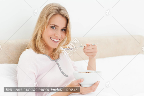 一個(gè)女人在床上拿著一勺麥片粥，在她手里拿著滿滿一碗麥片粥的時(shí)候，嘴里拿著勺子。