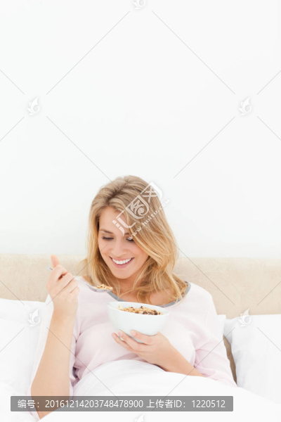 一個女人在床上垂直拍著一碗麥片粥；她微笑著看著麥片勺。