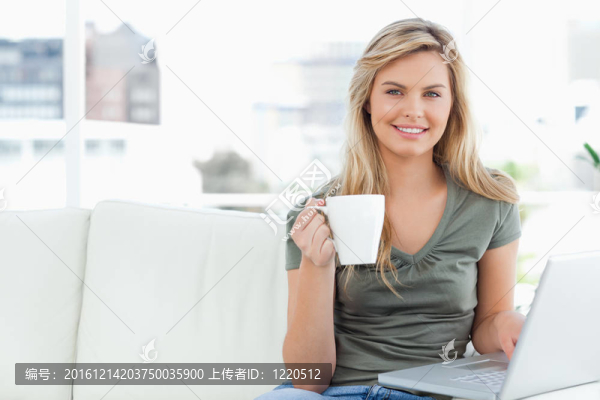 一名女子坐在沙發(fā)上，微笑，因?yàn)樗诖谒媲?；一個(gè)杯子在她的手和使用她的筆記本電腦。