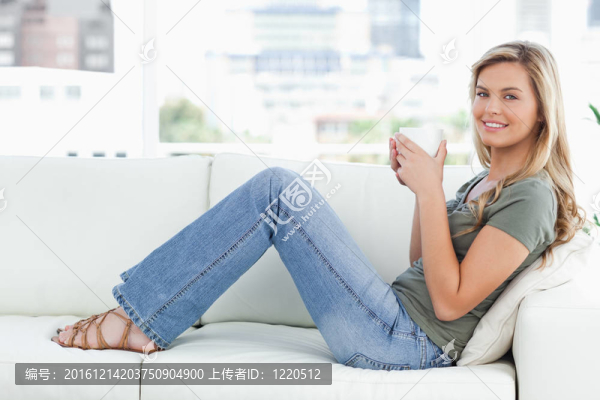 一個女人坐在沙發(fā)上，一邊向前看，一邊微笑著，手里拿著一個杯子。