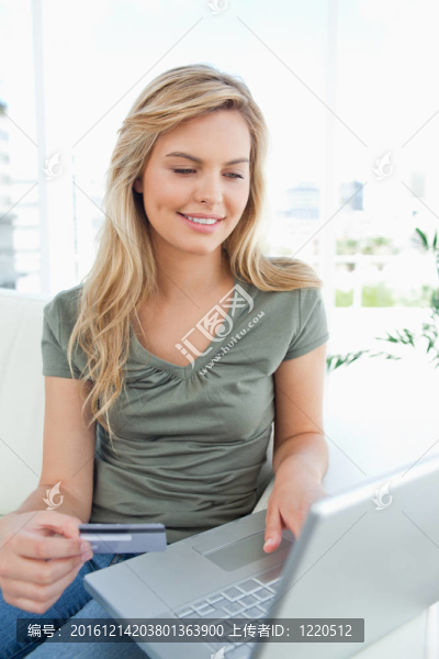 一個(gè)面帶微笑的女人一邊看著筆記本一邊拿著信用卡坐在沙發(fā)上。